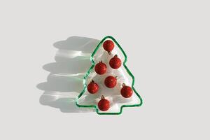 Plate as New Year tree with glittering red Christmas balls on white, Top view Christmas tree from green glowing glass. Natural Sunlight, sparkling holiday food, minimal xmas trendy still life photo