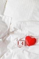 Valentine's Day concept, red paper valentine heart and 14 february holiday date on wooden calendar in bed on on white crumpled sheets. Minimal style flat lay photo, top view, copy space photo