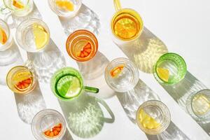 Summer still life top view pattern with colored glass cups of infused water with slices of fruit on white table. Glittering glasses at sunlight. Long harsh shadow. Summer vacation concept photo