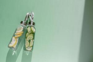 Glass bottles with lemon, cucumber water drink detox at sunlight on green colored background, copy space. Healthy infused water boost metabolism, weight loss. Aesthetics still life, refreshing drinks photo