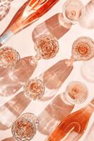Top view glasses and bottle of rose wine on peach color background with beautiful shadows at sunlight. Vintage wineglass minimal style pattern, closeup sparkling alcohol drink, summer drink concept photo