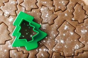 Cooking ginger cookies, Christmas New Year traditional cookies in shape of Christmas fir tree, raw rolled out dough with cut out dough figures, closeup, top view, green plastic pastry cutter on dough photo