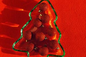 Green colored glass plate form as New Year tree with sparkling red Christmas balls instead of food, sunlight, shadow, star glare on red cloth background. Winter holiday dinner concept, minimal photo