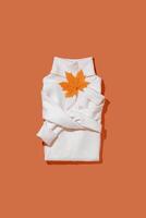 White warm folded sweater with autumn maple leaves on vivid orange color background, folded women jumper top view, minimal style trend autumn clothing concept, cozy clothes Aesthetic vertical Flatly photo