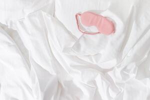 Pink eye mask for sleep on white bedclothes at home, minimal lifestyle aesthetic flat lay photo. Top view Female sleeping mask for best sleepers, for travel, comfort relaxation. Rest well concept. photo