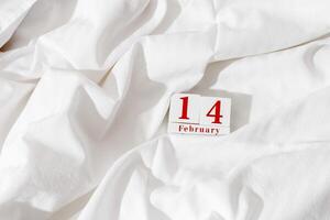 14 february background, holiday date on calendar in bed with white cotton sheets. Valentine's Day minimal concept, aesthetic lifestyle photo. Wooden calendar with red text, festive romance date photo