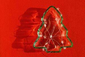 Plate as New Year tree with Glowing light stars garland on red textile, Christmas tree from transparent green glowing glass glare. Sunlight and shadow, sparkling holiday dish, xmas still life photo