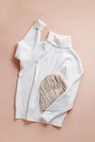 White turtleneck or sweater and knitted hat, casual garment, comfort and style warm clothes. Soft pullover and wool hat on beige pink color background, minimal fashion clothing, top view photo
