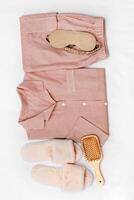 Top view of folded woman pink pajama, beige sleep eye mask, hair brush, soft home slippers on white. Minimal style accessories and cozy pyjamas. Fashion sleepwear, calm lifestyle, home aesthetic photo