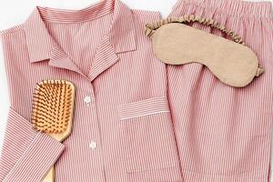 Flat lay with pink Pajama, beige sleep eye mask, hair brush on white sheet. Top view pastel pink color woman home sleepwear and accessories on bedclothes. Cozy pyjamas of shirt and shorts for sleep photo