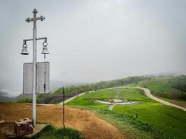 Georgia, Didgori, Sakartvelo. Metal bells and a cross rise from a misty hilltop, Metal bells and a cross rise from a misty hilltop, marking a tranquil spiritual monument along a winding rural pathway photo