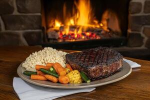 A steak and vegetables on a plate in front of a fireplace photo
