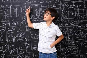 A young boy in glasses standing in front of a blackboard with math formulas written on it photo