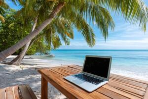 Laptop computer on wooden table at tropical beach under palm trees with turquoise water photo