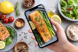 Food blogger taking photo of delicious grilled salmon, asparagus, and lemon with smartphone