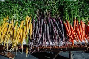 Freshly harvested heirloom carrots in yellow, purple, and orange, arranged in rows with green tops still attached. Displayed at an outdoor market table, ideal for content on healthy eating photo
