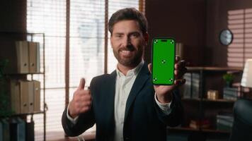 Smiling happy middle aged business man showing green screen mobile phone like gesture thumb up holding smartphone chroma key display success achieve mature businessman male looking at camera in office video