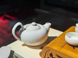 White ceramic teapot on light surface. Round teapot with curved handle and short spout next to bamboo tea tray. photo