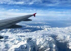stunning aerial view of rugged snow-covered mountain peaks stretching into the horizon, viewed from an airplane window with the wing partially visible, under vivid blue sky with high-altitude clouds photo