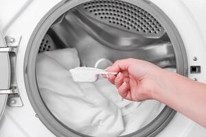 hand holding measuring spoon with washing powder against background of washing machine with white laundry photo