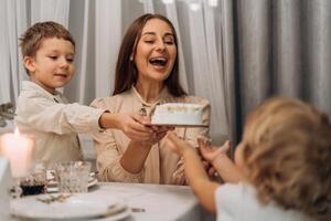 Happy mother laughing while holding cake together with smiling older child and reaching hands of toddler during warm joyful family gathering in cozy home setting full of love connection and sweet photo