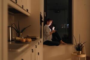 Woman sits in windowlight alone at night looking at glowing smartphone representing blue light at night digital intimacy and emotional solitude during evening scrolling routine in modern lifestyle photo