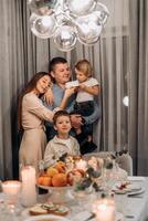 Smiling family gathered festive dinner table father holds toddler eating white cake mother lovingly supports child and older sibling stands nearby capturing heartwarming moment connection celebration photo