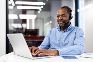 Mature african american businessman in modern office setting using laptop and headset. Displaying professionalism, communication skills, and confidence while typing and engaging with clients. photo
