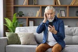 An elderly man sits on a sofa, looking confused and worried as he reads a message on his mobile phone, portraying a moment of concern. photo