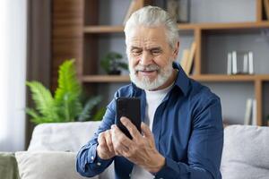 Cheerful elderly man with a gray beard enjoying technology, texting or browsing on his smartphone in a cozy living room setting. photo