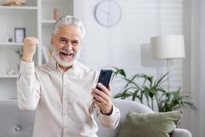 Happy gray-haired senior man is at home, sitting on the couch, looking at the screen of the phone he is holding and rejoicing in success, showing a victory gesture with his hand. photo