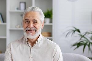 Close-up portrait of a smiling gray-haired senior man at home, sitting on a sofa in a white shirt and with a beard, smiling at the camera. photo