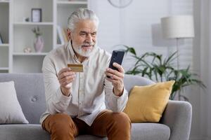 Smiling senior gray-haired man using mobile phone and credit card at home and sitting on sofa. photo