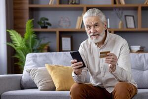Portrait of a smiling senior man at home, sitting on the couch and looking at the camera. holds a credit card and mobile phone. photo