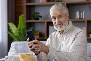 Portrait of senior man, grandfather at home, sitting on sofa and using phone, smiling and looking at camera. photo