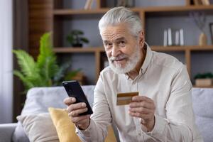 Close-up portrait of a handsome senior man with gray hair in a shirt sitting on a sofa at home, using a phone and a credit card, looking at the camera with a smile. photo