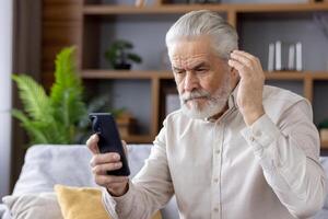 Elderly man with gray hair and beard looking at his smartphone with a confused expression while sitting on a comfortable couch at home. photo