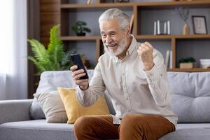 Happy senior man enjoying success showing victory gesture with hand, sitting on sofa at home and using mobile phone. photo