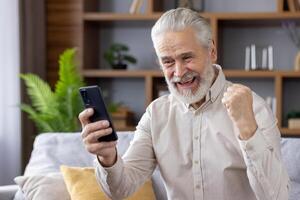 Happy senior man grandpa enjoying success, sitting on sofa at home, looking at mobile phone screen and showing victory gesture with hand. photo