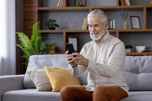 Senior man sitting on a couch in a cozy living room, using a smartphone and smiling. Concept of technology, communication, and leisure. photo