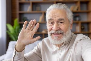 Close-up photo of an older gray-haired man with a beard talking on the phone camera, taking a selfie at home, greeting and waving.
