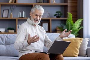 Gray-haired old smiling man is having a call on a laptop, sitting on the sofa at home and talking remotely. photo