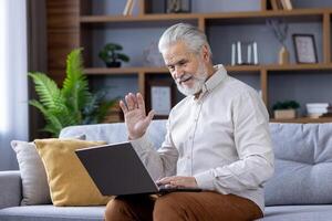 Smiling gray-haired senior man sitting at home on sofa with laptop and talking on call to camera, waving and greeting. photo