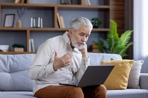 Upset senior gray-haired man talking on call with family and bank, sitting on sofa and solving financial problems and questions. photo