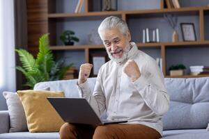 Senior gray-haired man sitting at home on the sofa, holding a laptop on his lap and looking happy at the camera of the communication, showing a victory yes gesture with his hands. photo