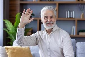 Portrait of an older gray-haired smiling man sitting at home on the sofa in front of the camera, greeting and waving. photo