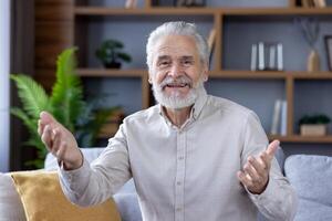 Senior gray-haired man sitting on the sofa at home and talking on a call to the camera, gesturing with his hands and smiling. photo