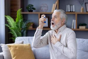 An elderly gray-haired man suffers from asthma. He sits at home on the couch and uses an inhaler. photo