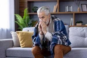 A sick gray-haired man is sitting at home on the sofa covered with a warm blanket, blowing his nose from a cold, suffering from colds and the flu. photo