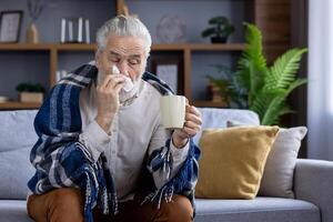 An elderly man is not feeling well, sitting at home on the sofa covered with a blanket, holding a cup with a drink in his hands and wiping his nose with a napkin from a runny nose. photo